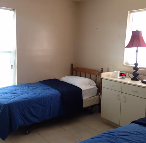 A small bedroom with two single beds covered with blue blankets. One bed is positioned against a beige wall with a wooden headboard. Next to the bed is a white cabinet with drawers and a red lamp on top. A digital clock displaying 4:22 and a roll of toilet paper are also on the cabinet. There are two windows allowing natural light into the room.