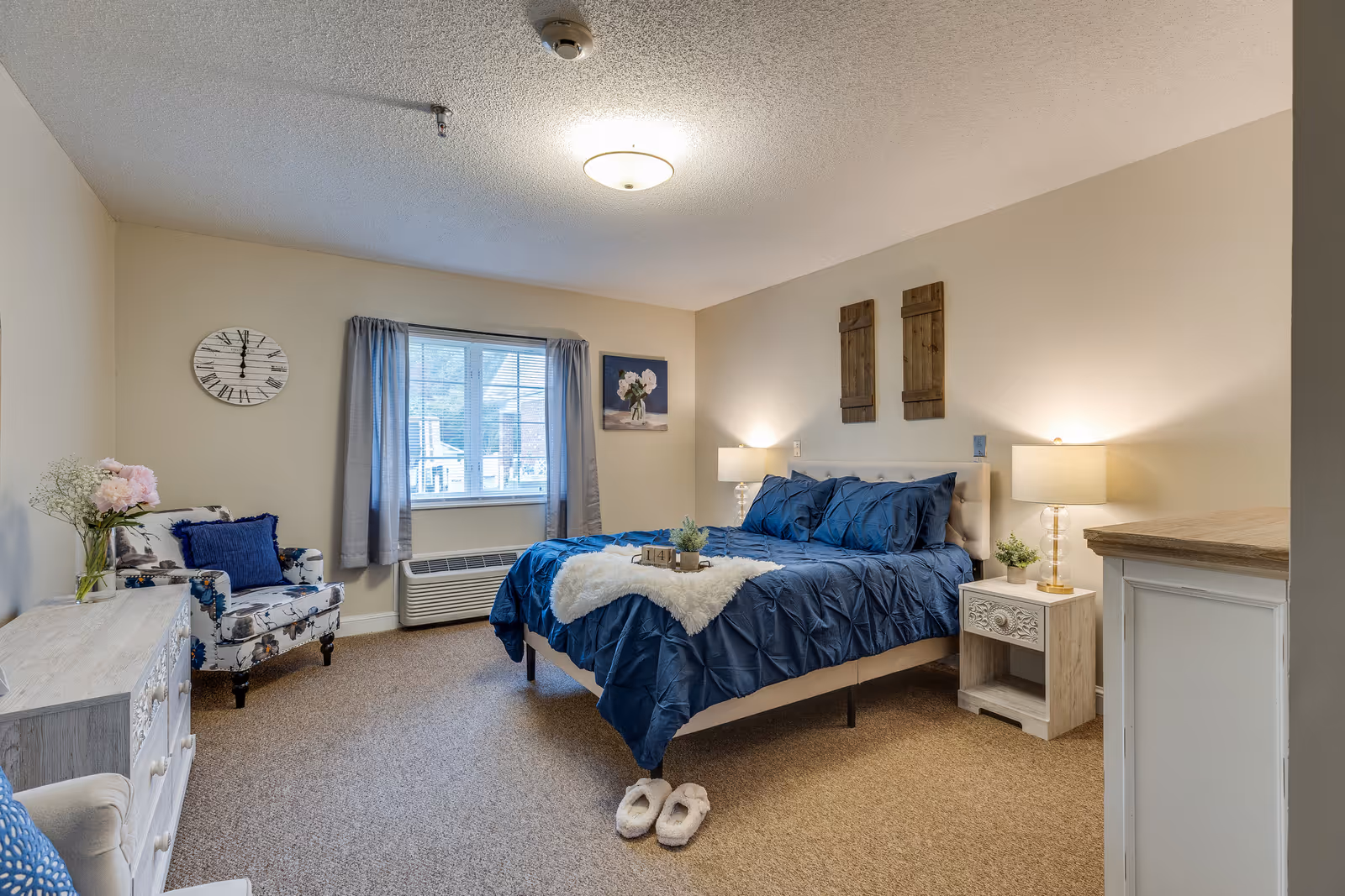 A cozy bedroom in an assisted living facility featuring a bed with blue bedding and a white throw, two bedside tables with lamps, a floral armchair, a window with gray curtains, a wall clock, and decorative wall art. The room has beige walls and carpeted flooring.
