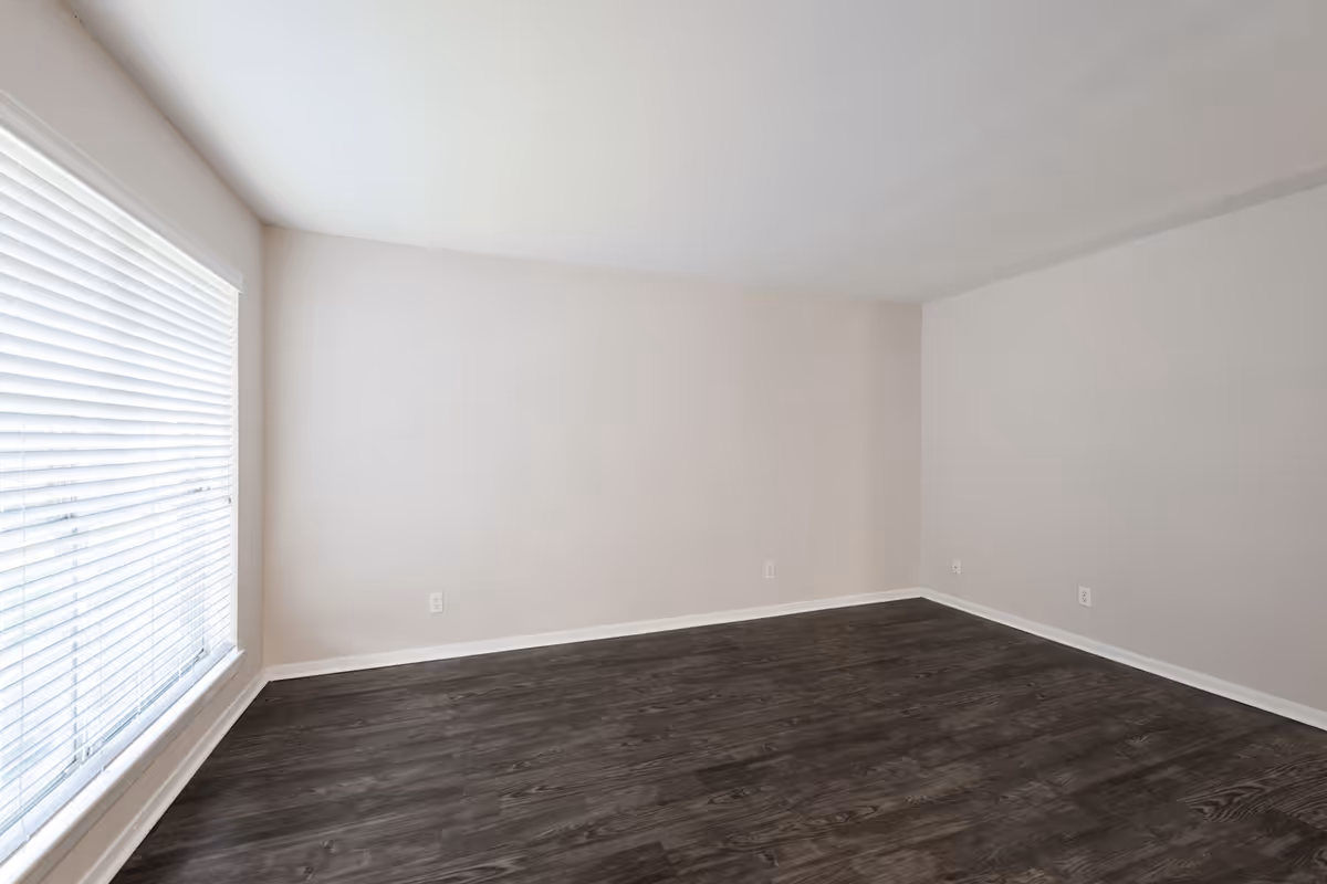 Empty room with light beige walls, dark wood flooring, and a large window with white blinds letting in natural light.