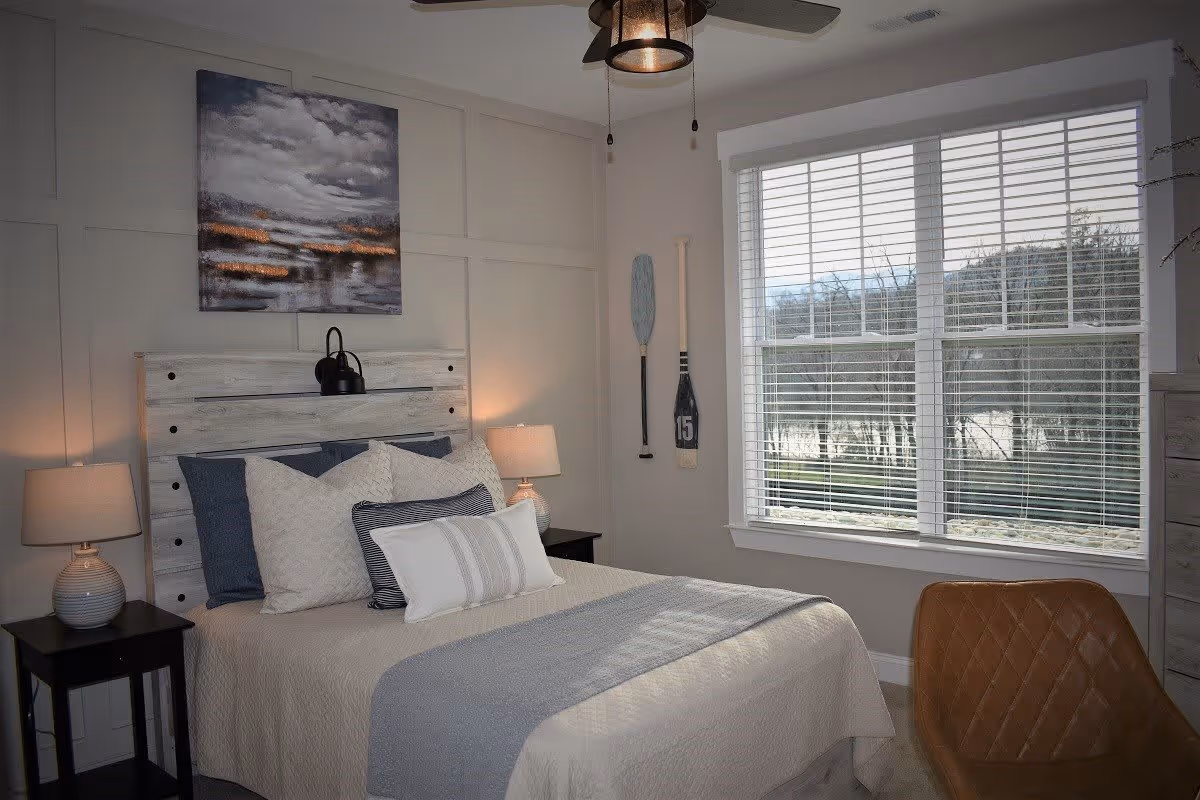 A cozy bedroom with a bed featuring a light-colored quilt and multiple pillows in shades of white and blue. The headboard is rustic wood with a black reading lamp attached. On either side of the bed are black nightstands with matching lamps. Above the bed hangs a painting of a cloudy sky over a body of water. Two decorative oars are mounted on the wall next to a large window with white blinds, through which trees and a body of water are visible. A brown leather chair is positioned near the window.
