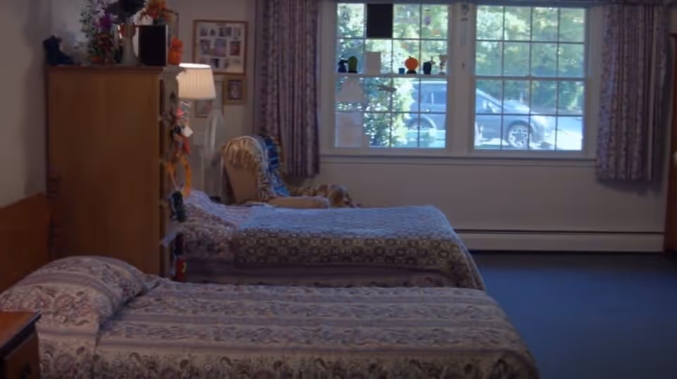 A bedroom with two single beds covered in patterned bedspreads. There is a wooden dresser with a lamp and decorative items on top. A window with curtains lets in natural light, and a chair with a blanket is placed near the window.