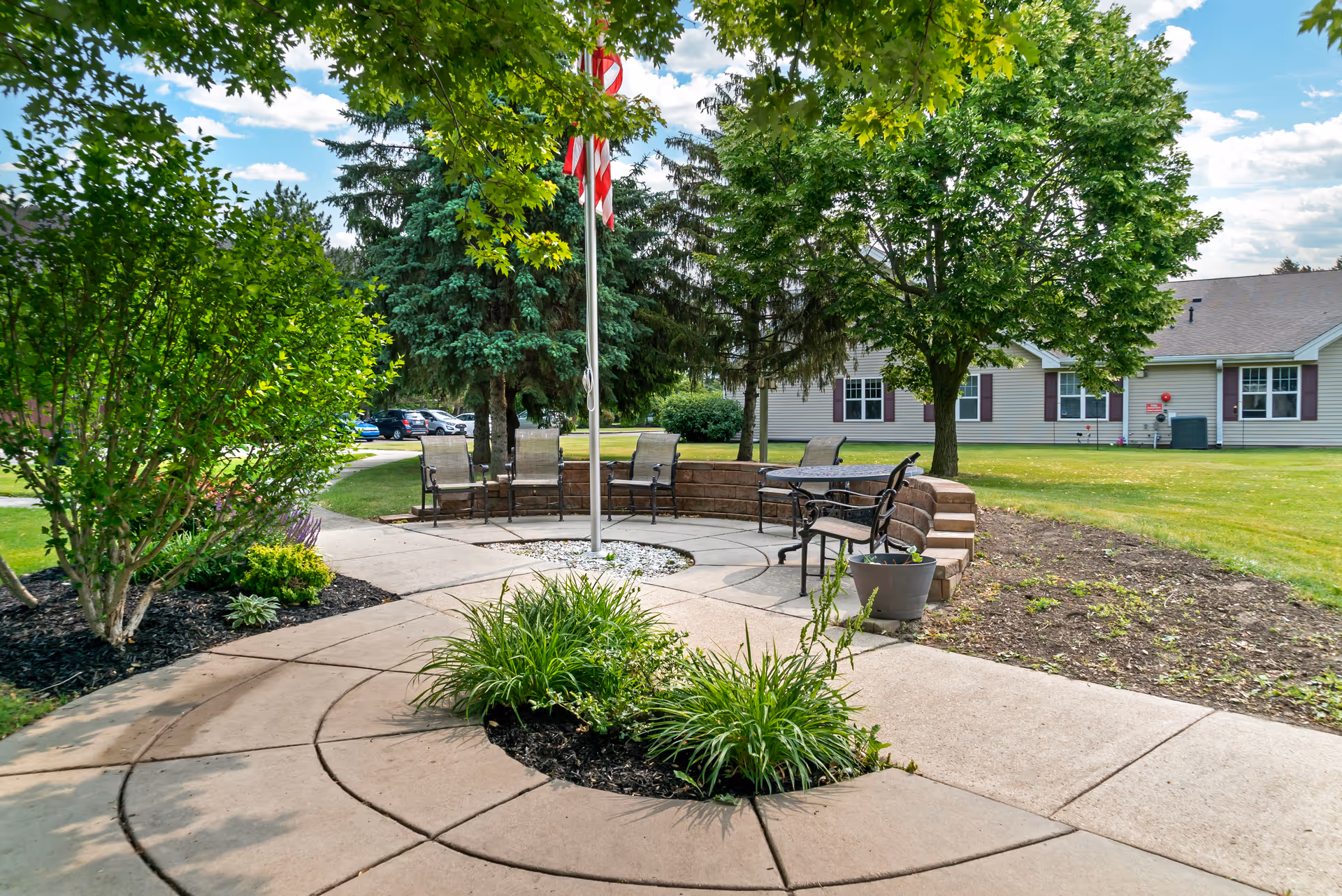 Sunlit courtyard with a circular paved seating area around a flagpole, benches and patio furniture in front of a single-story building.