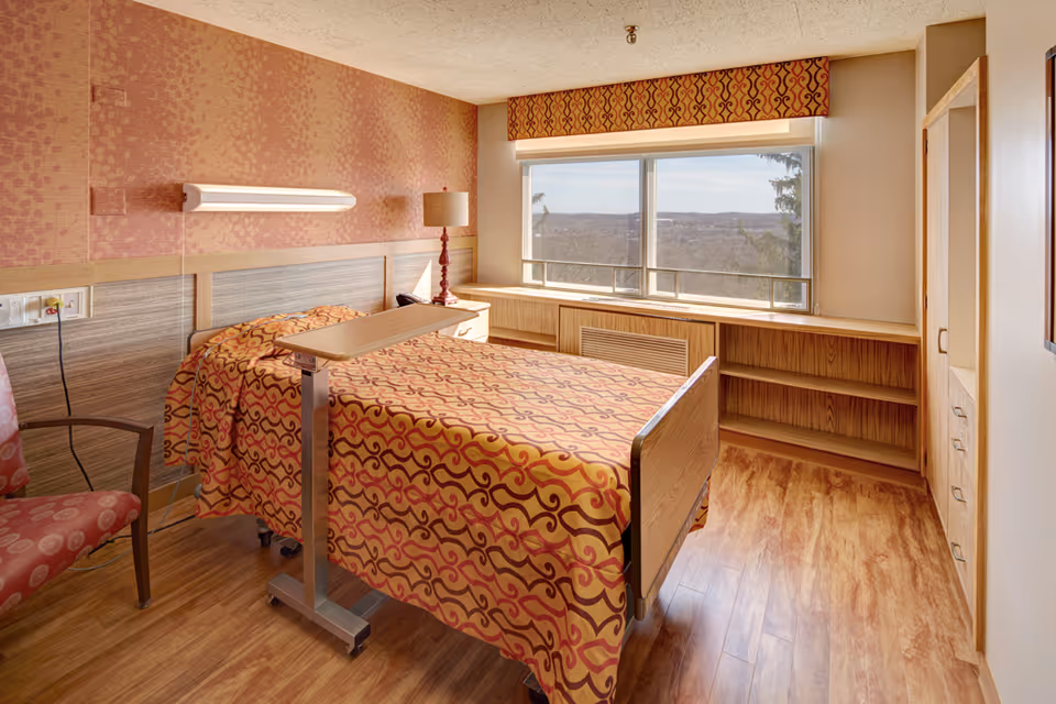 A bright senior facility bedroom with an adjustable hospital bed, overbed table, patterned bedding, armchair, lamp and a large window with a view.