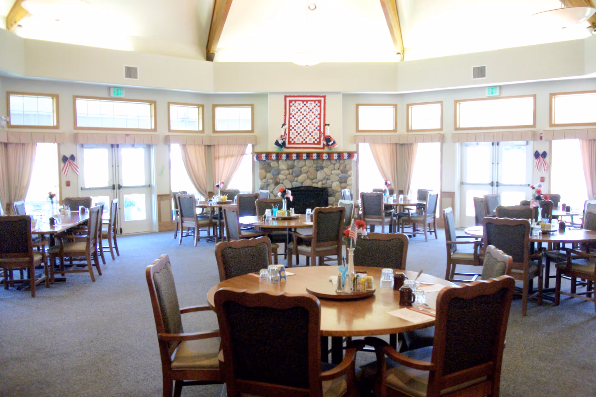 A spacious dining room with multiple round and rectangular tables surrounded by wooden chairs with cushioned seats. Each table is set with glasses, condiments, and small flower vases. The room features large windows with beige curtains, a stone fireplace with a quilt hanging above it, and a high ceiling with wooden beams.