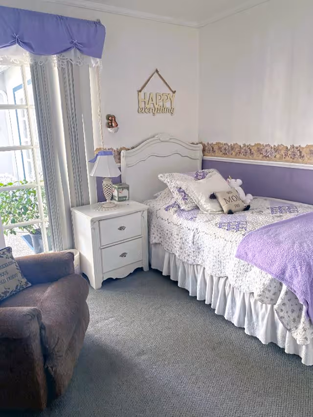 A cozy bedroom with a single bed covered in a white and purple quilt, several pillows including one with the text 'Be My', and a small stuffed animal. Next to the bed is a white nightstand with a lamp and a small decorative item. A brown armchair with a pillow is positioned near a window with vertical blinds and a purple valance. The walls are decorated with a floral border and a hanging sign that reads 'HAPPY everything'.