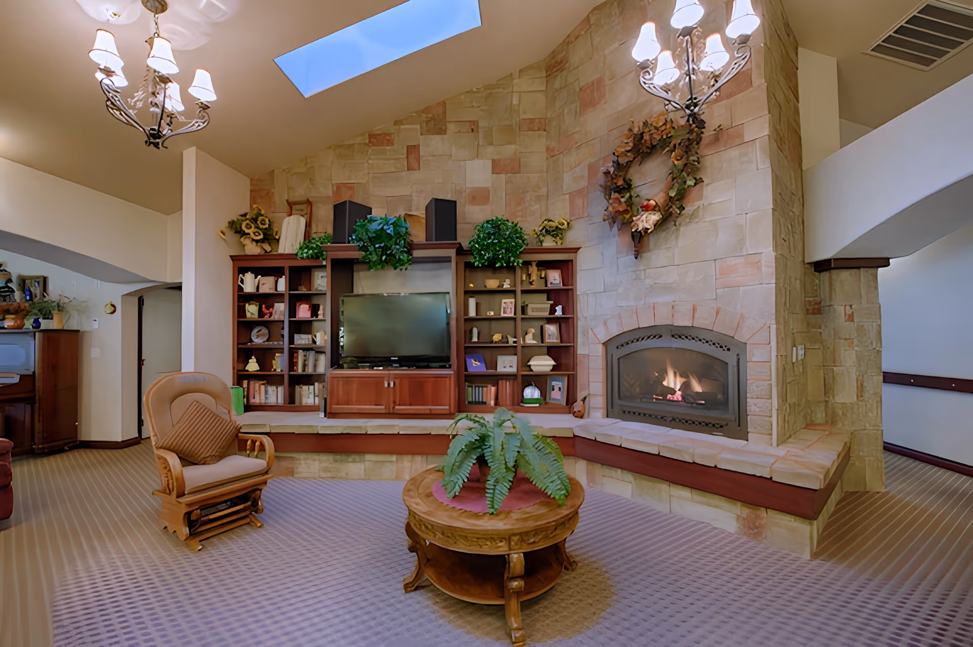 A cozy living room area with a stone fireplace on the right side, a wooden entertainment center with a TV and shelves filled with books and decorative items, a wooden rocking chair with cushions, a round wooden coffee table with a green fern plant, and two chandeliers hanging from the ceiling with a skylight above.