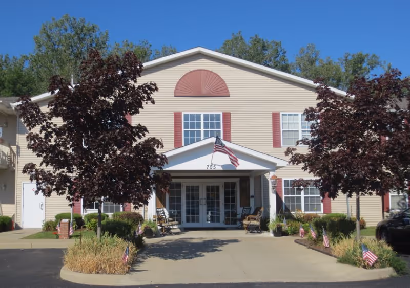 Front exterior view of Sandra Lane Senior Apartments building with beige siding, red shutters, and an American flag above the entrance. There are two trees and several small American flags lining the walkway leading to the entrance.