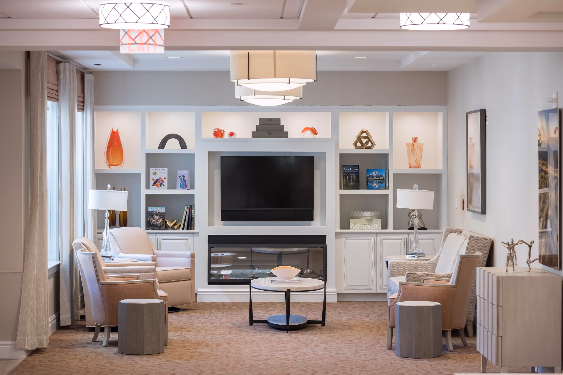 A cozy and well-lit living room area with a wall-mounted flat-screen TV above a modern electric fireplace. The room features built-in white shelves with decorative items and books, two pairs of beige armchairs with side tables and lamps, and a round coffee table in the center. Large windows with curtains allow natural light to enter the space.