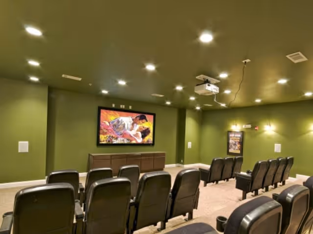A small movie theater room with rows of black cushioned seats facing a large screen mounted on a green wall. The ceiling has recessed lighting and a projector is hanging from it. There is a framed movie poster on the right wall.
