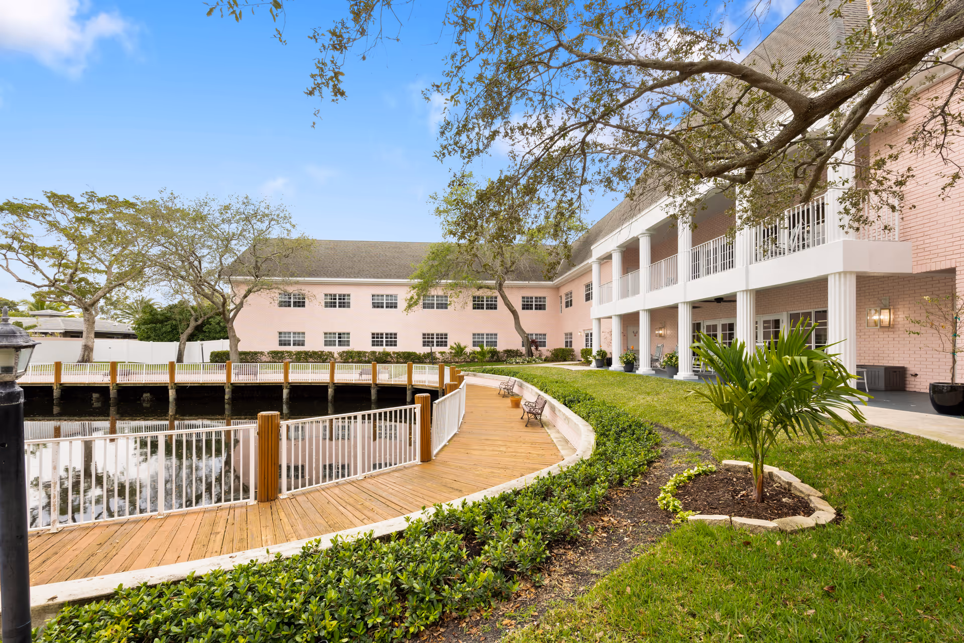 Exterior view of a pink two-story assisted living building with a riverside wooden boardwalk, landscaped lawn, and trees under a blue sky.