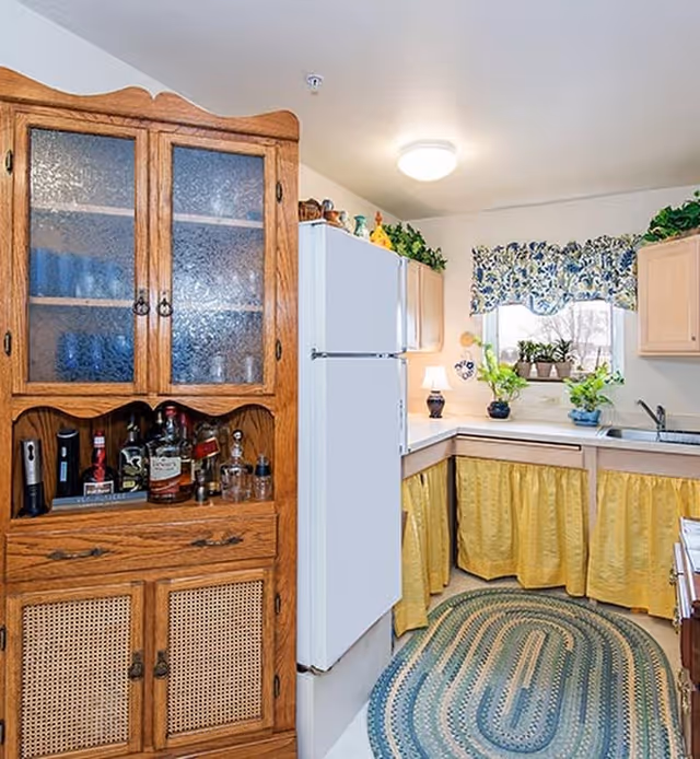 A cozy kitchen with a white refrigerator, wooden cabinets with yellow curtains underneath the countertops, a small lamp on the counter, potted plants on the windowsill, and a wooden cabinet with glass doors containing bottles and glassware. A blue and green braided rug is on the floor.