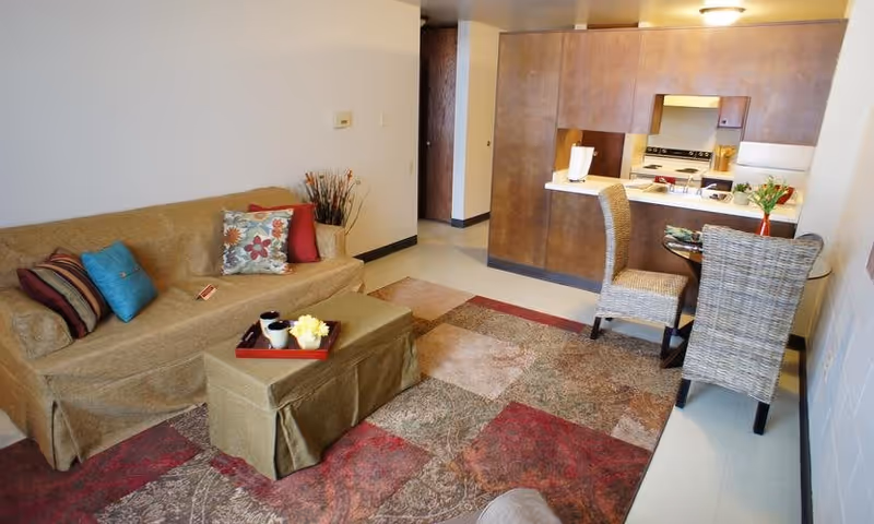 Interior view of a senior living facility apartment featuring a beige sofa with colorful cushions, a matching ottoman with a tray holding cups and a flower, a patterned area rug, a small dining table with two wicker chairs, and a compact kitchen area with wooden cabinets and a stove.