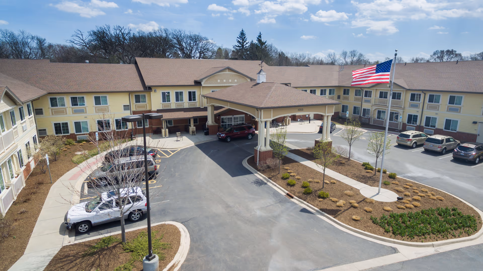 Exterior view of a large, two-story assisted living and memory care facility with a covered entrance driveway. Several cars are parked in the parking lot, and an American flag is flying on a flagpole in the landscaped area near the entrance. Trees and shrubs surround the building under a partly cloudy sky.