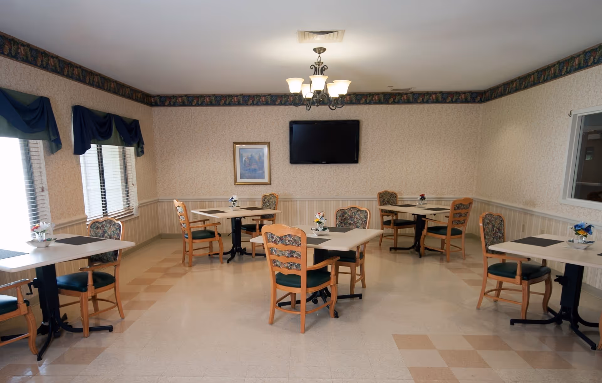 A small dining room with several square tables, each surrounded by wooden chairs with floral-patterned cushions. The room has beige wallpaper with a floral border near the ceiling, a chandelier light fixture, a wall-mounted flat screen TV, and a framed picture on the wall. Windows with blue and green valances allow natural light into the room.