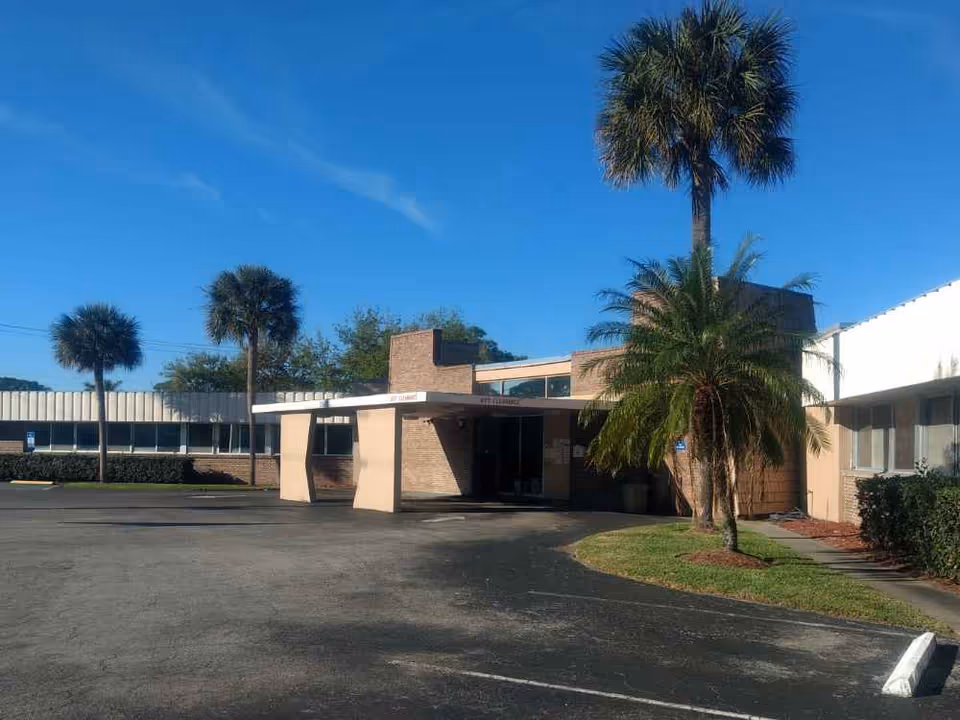 Front entrance of a low-rise brick building with palm trees and a parking lot under a clear blue sky.