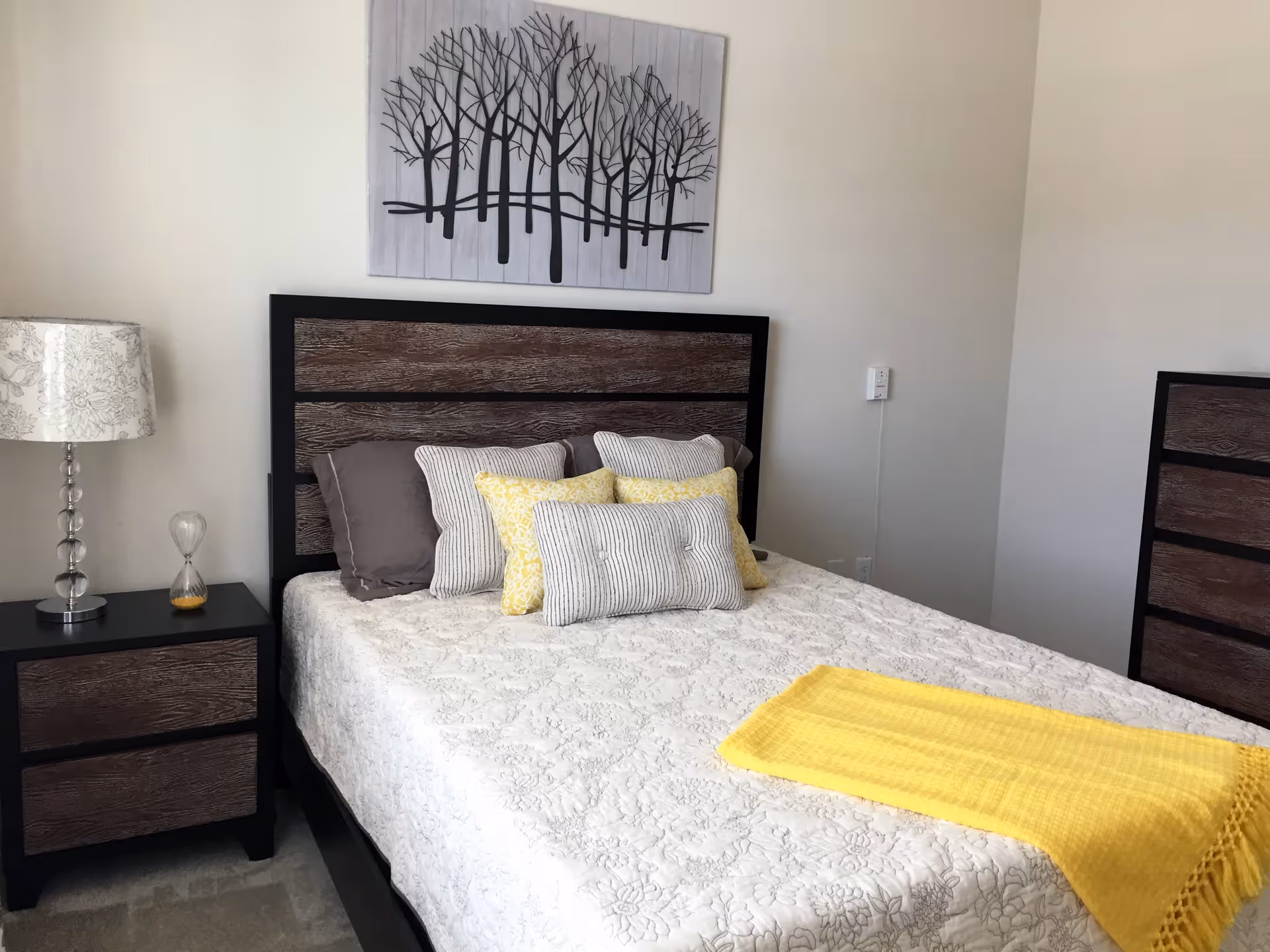 A neatly made bed with a wooden headboard and multiple pillows in shades of gray and yellow. A yellow throw blanket is draped across the foot of the bed. To the left of the bed is a nightstand with a decorative lamp and an hourglass. Above the bed hangs a piece of wall art depicting a group of leafless trees. The room has light-colored walls and a dresser matching the bed's wood finish is partially visible on the right.