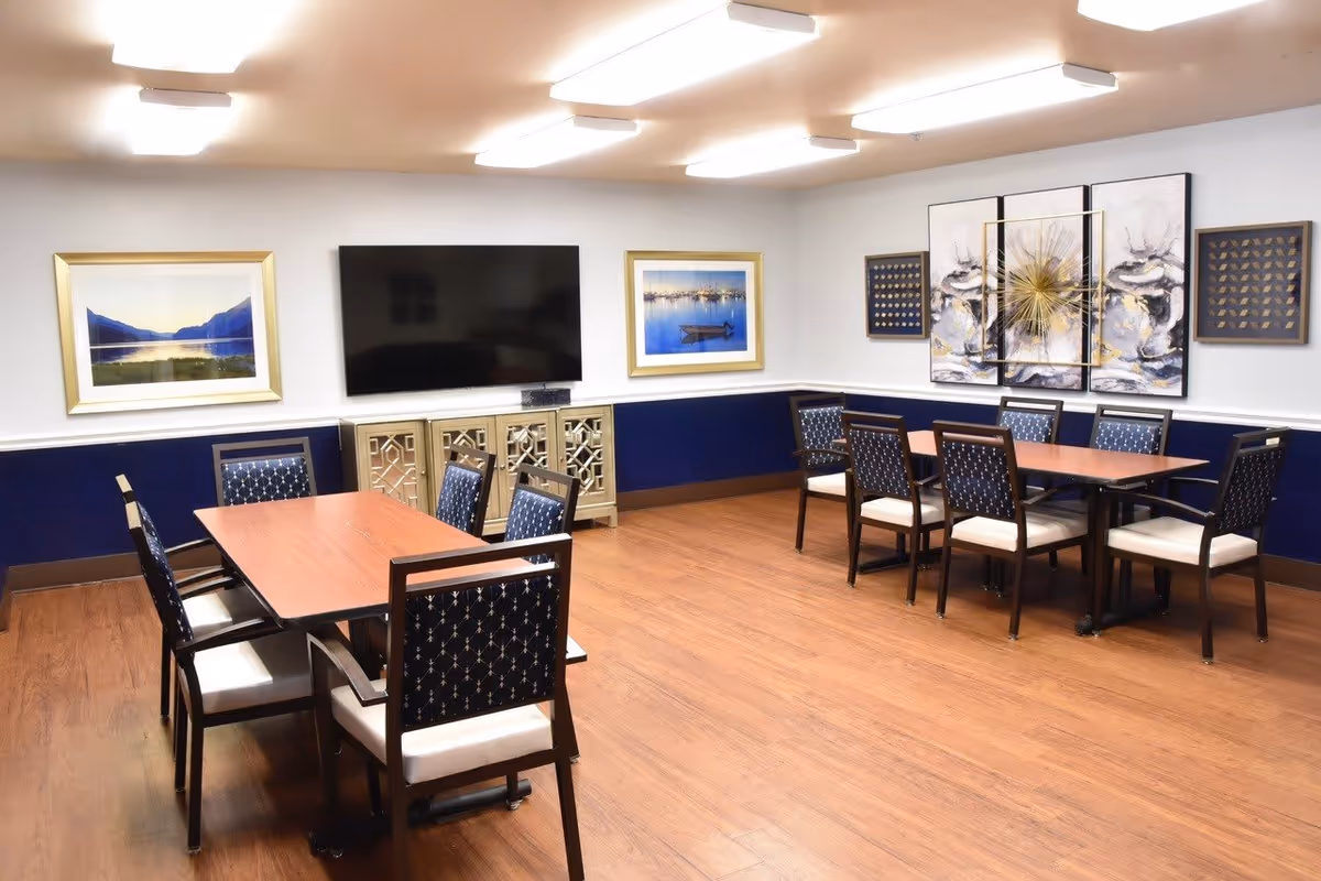 A well-lit room with two wooden tables surrounded by chairs with dark blue patterned upholstery and white cushions. The room has wood flooring, white walls with a dark blue lower section, and several framed artworks including landscape photos and abstract paintings. A large flat-screen TV is mounted on the wall above a decorative cabinet.