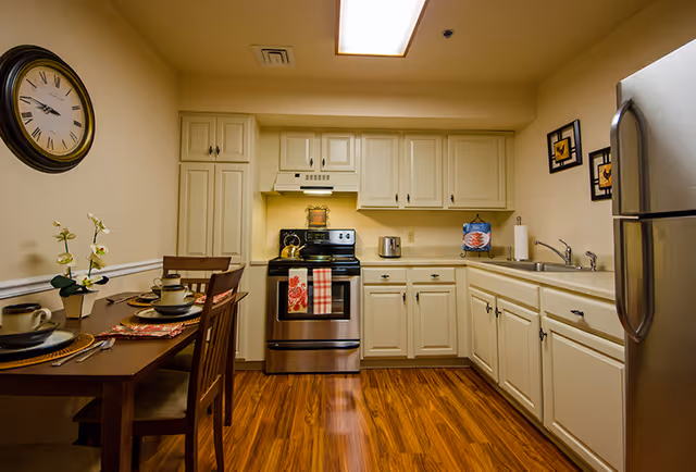 A clean and well-lit kitchen with white cabinets, a stainless steel stove and refrigerator, a double sink, and a wooden dining table set for four with plates, cups, and cutlery. A large wall clock is visible on the left wall, and two framed pictures hang on the right wall.