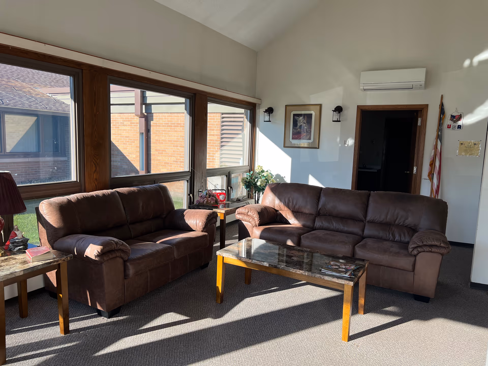 Sunlit sitting area with two brown leather sofas, a marble-top coffee table, large windows, and an American flag in the corner.