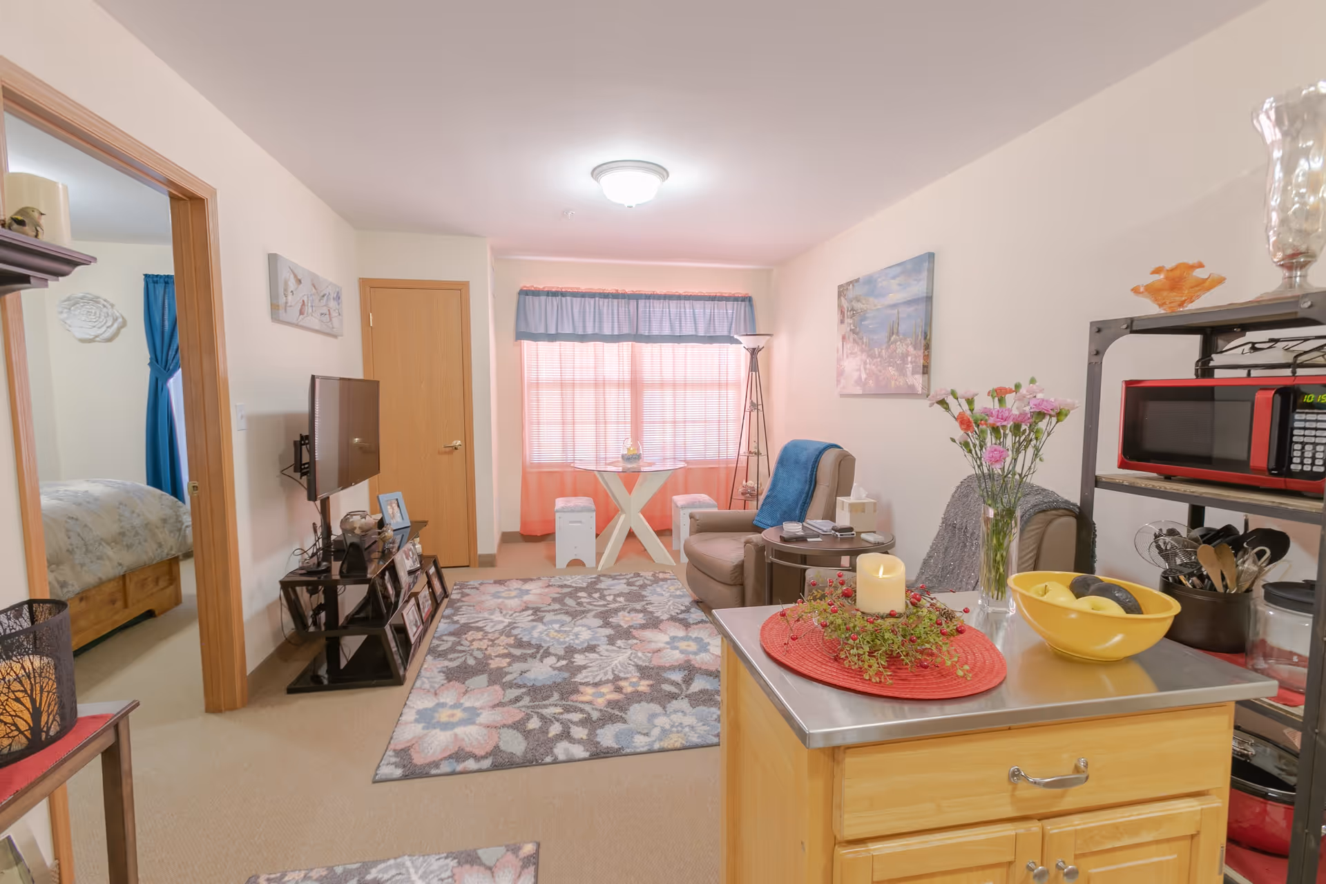 A cozy living room area in a senior living facility with a floral rug, a beige armchair with a blue throw, a small round side table with a tissue box and remote controls, and a small dining table with two stools near a window with blue and pink curtains. A flat-screen TV is mounted on the wall opposite the armchair. A kitchen island with a yellow bowl of fruit, a candle centerpiece, and a vase of flowers is visible in the foreground. A black metal shelving unit with a red microwave and kitchen utensils is on the right side. A bedroom with a bed and blue curtains is visible through an open door on the left.