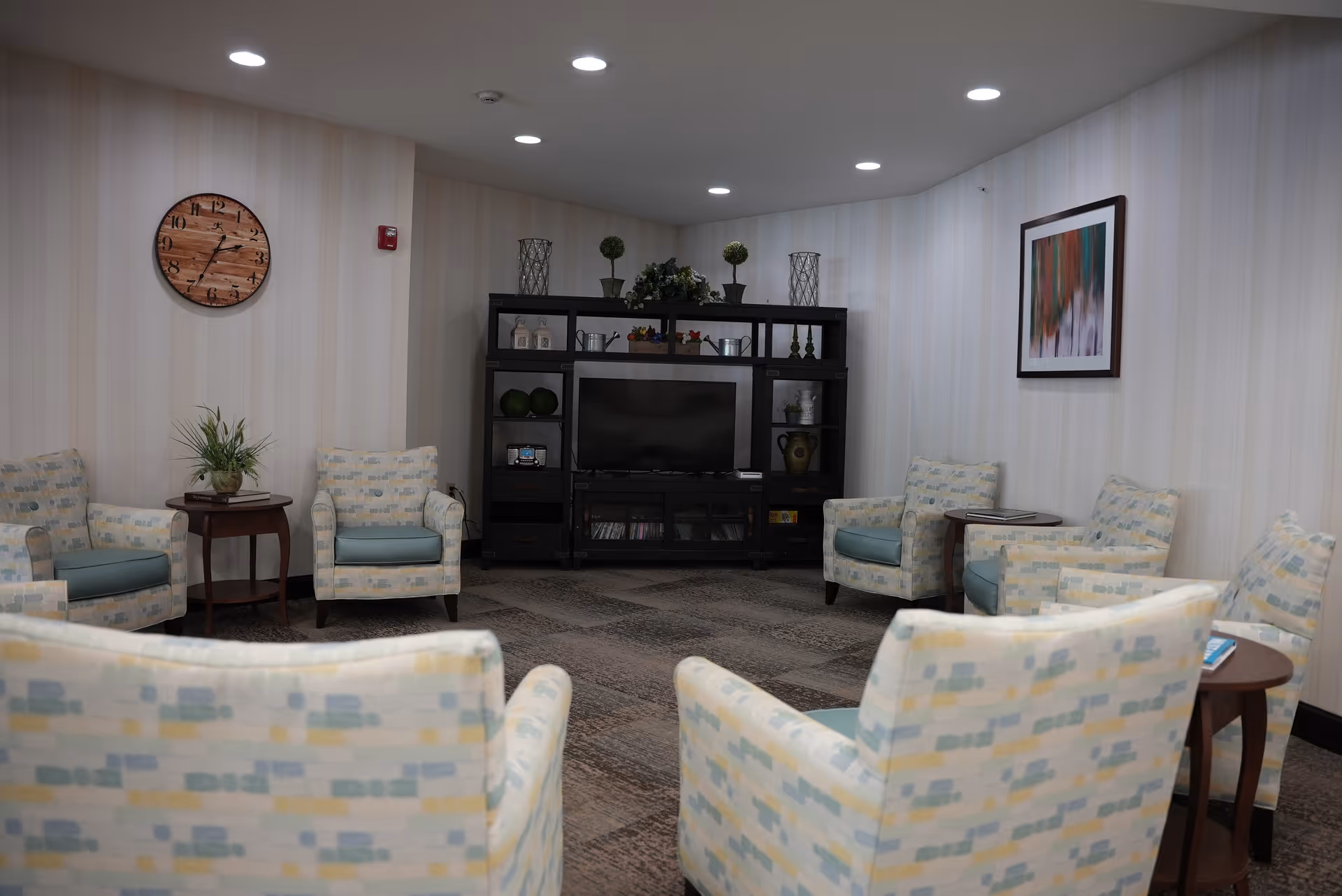 A cozy living room area with patterned armchairs arranged in a circle around small wooden side tables. A black entertainment center with a flat-screen TV and decorative items is against the far wall. The walls have light-colored vertical stripes, and a wooden clock and framed abstract artwork are hung on the walls. The room is softly lit with recessed ceiling lights.
