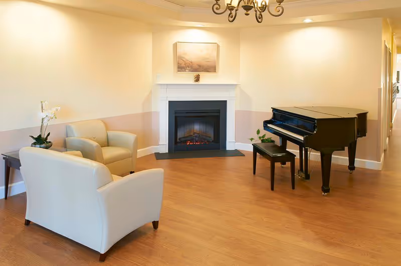 A cozy living room area with light beige walls and wooden flooring featuring a black grand piano with a matching bench, a white fireplace with a painting above it, two beige armchairs, a small side table with a white orchid plant, and a chandelier hanging from the ceiling.