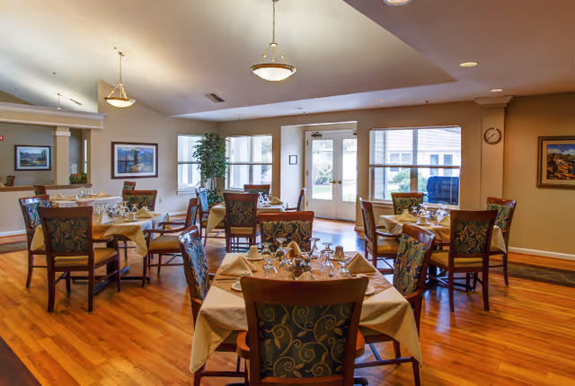 A dining room with several tables covered with beige tablecloths, each set with glassware, napkins, and silverware. The room has wooden floors, large windows letting in natural light, framed artwork on the walls, and ceiling light fixtures. There is a door leading outside and some greenery near the windows.