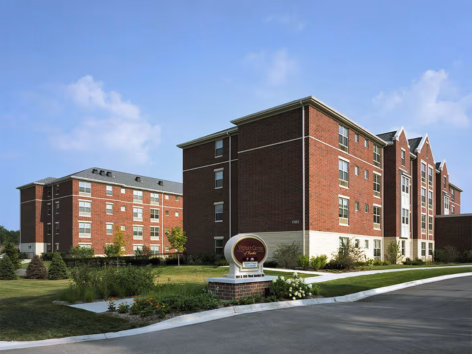 Brick multi-story senior living facility with landscaped grounds and a circular Victory Centre sign by the driveway.