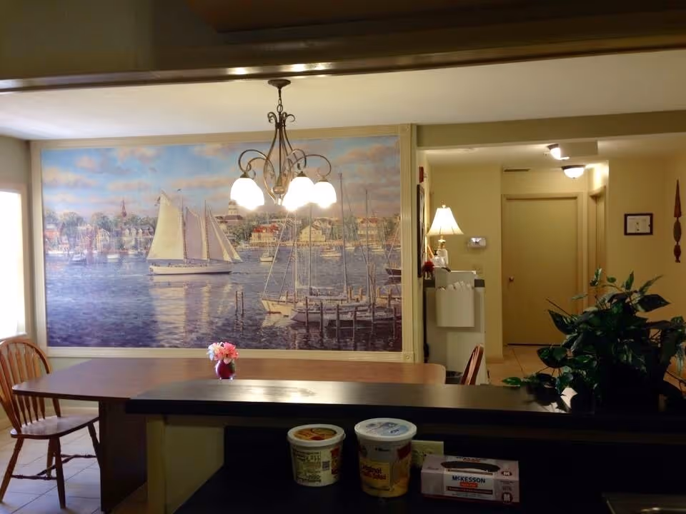 Interior view of a dining area with a wooden table and chairs. A large mural of sailboats on water with a town in the background covers one wall. A chandelier with five lights hangs above the table. There is a small vase with pink flowers on the table. In the background, a hallway with closed doors and a lamp on a small table is visible. A green plant is on the right side near the counter, which has some containers and a box of McKesson gloves.
