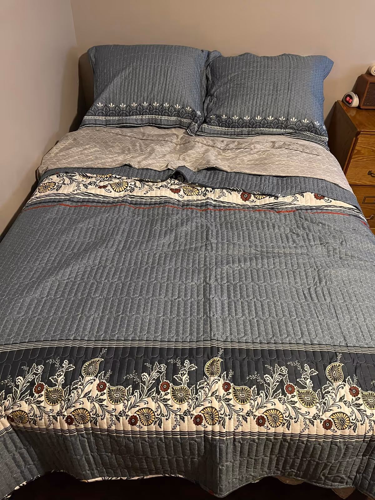 A neatly made bed with a quilted bedspread and matching pillows in shades of blue and beige with floral patterns. There is a wooden nightstand to the right of the bed with a digital alarm clock and a small lamp.