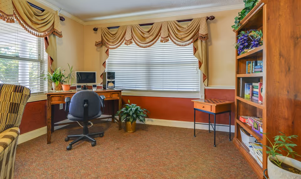 A cozy room with a wooden desk holding a computer and speakers, a black office chair, a small table with a chessboard, a bookshelf filled with books and games, several potted plants, and windows with beige and gold drapes.