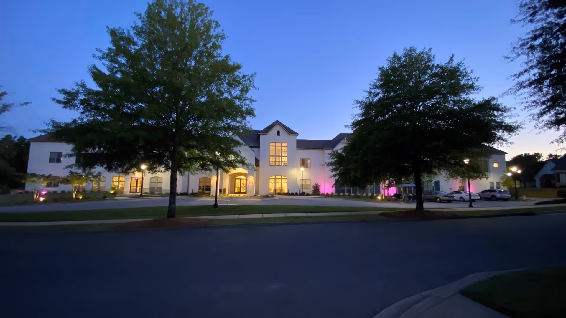 Front exterior of a two-story senior living building at dusk, illuminated and set behind trees with cars parked nearby.
