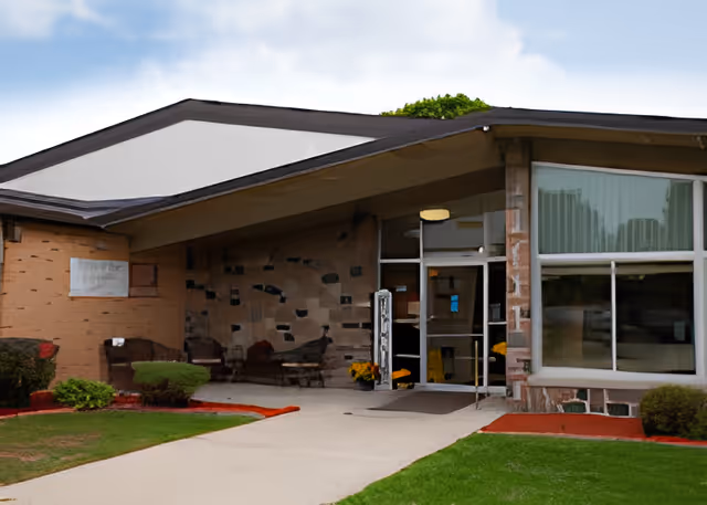 Entrance of a single-story senior care building with a sloped roof, covered walkway, glass double doors and large front windows.