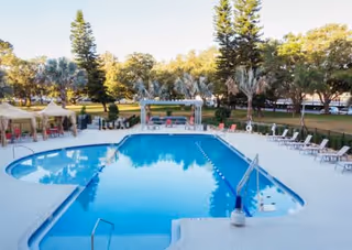 Outdoor swimming pool surrounded by lounge chairs and shaded seating areas with trees and greenery in the background under a clear sky.