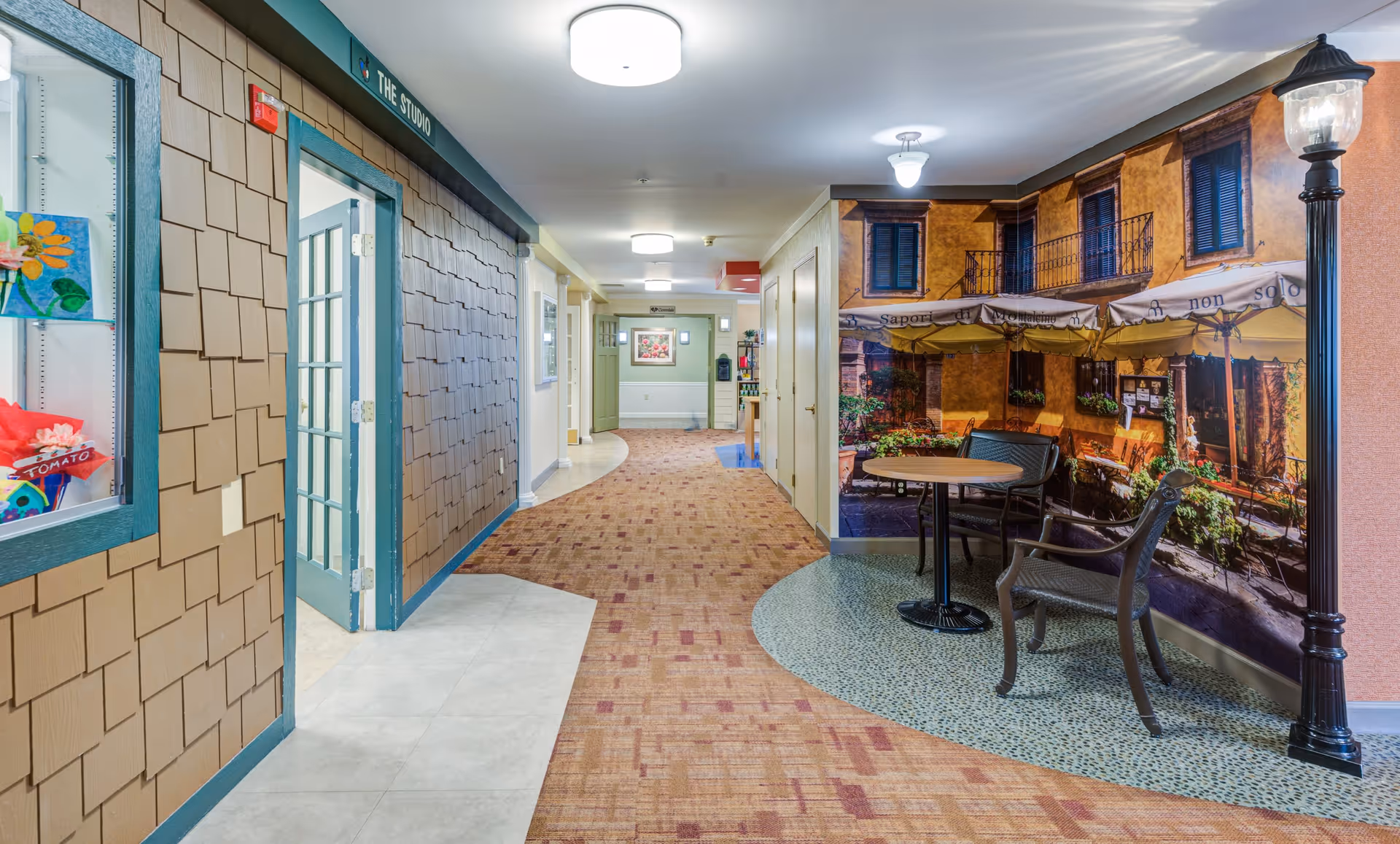 Interior hallway of a senior living facility with a carpeted floor and beige walls. On the right side, there is a small seating area with a round table and two chairs, set against a wall mural depicting an outdoor café scene with yellow buildings and umbrellas. On the left side, there is a door labeled 'The Studio' and a display case with colorful artwork. The hallway is well-lit with ceiling lights.