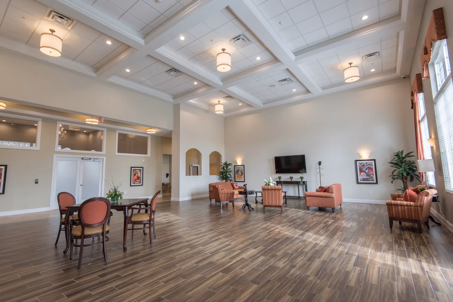 A spacious, well-lit common area with high coffered ceilings and wooden flooring. The room features a seating area with several upholstered chairs and sofas arranged around a coffee table, a wall-mounted flat-screen TV, and a small dining table with four chairs. Large windows with red valances allow natural light to fill the space, and there are framed artworks and potted plants decorating the walls and corners.