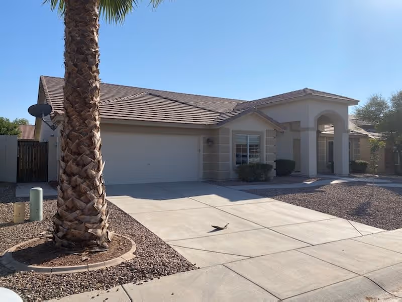 Single-story residential building with a tiled roof, a two-car garage, and a driveway. There is a palm tree in the front yard surrounded by gravel landscaping. The sky is clear and blue.