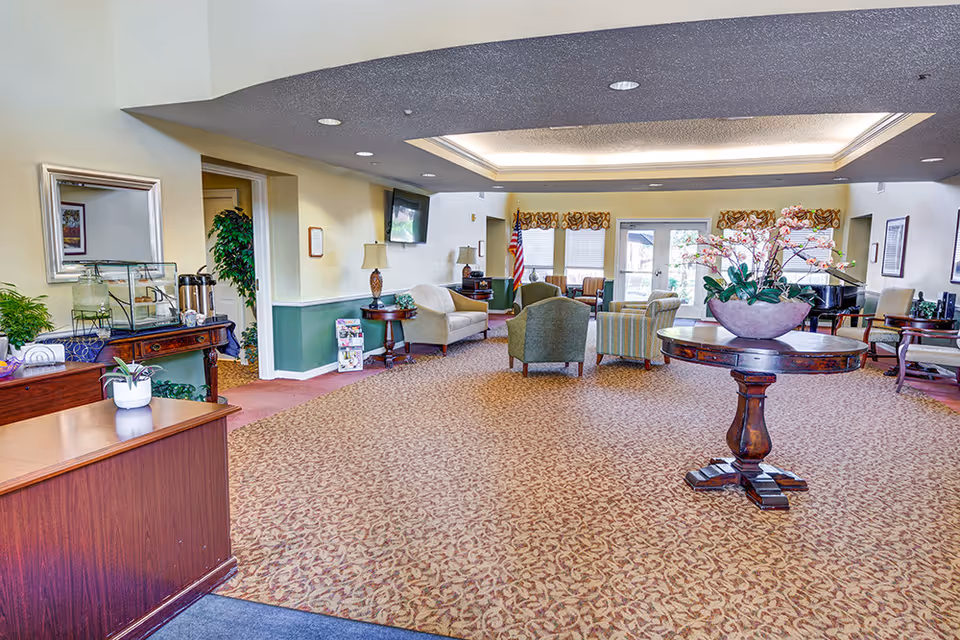 Spacious communal lounge in a senior living facility with armchairs, sofas, a round table with flowers, and a reception desk.