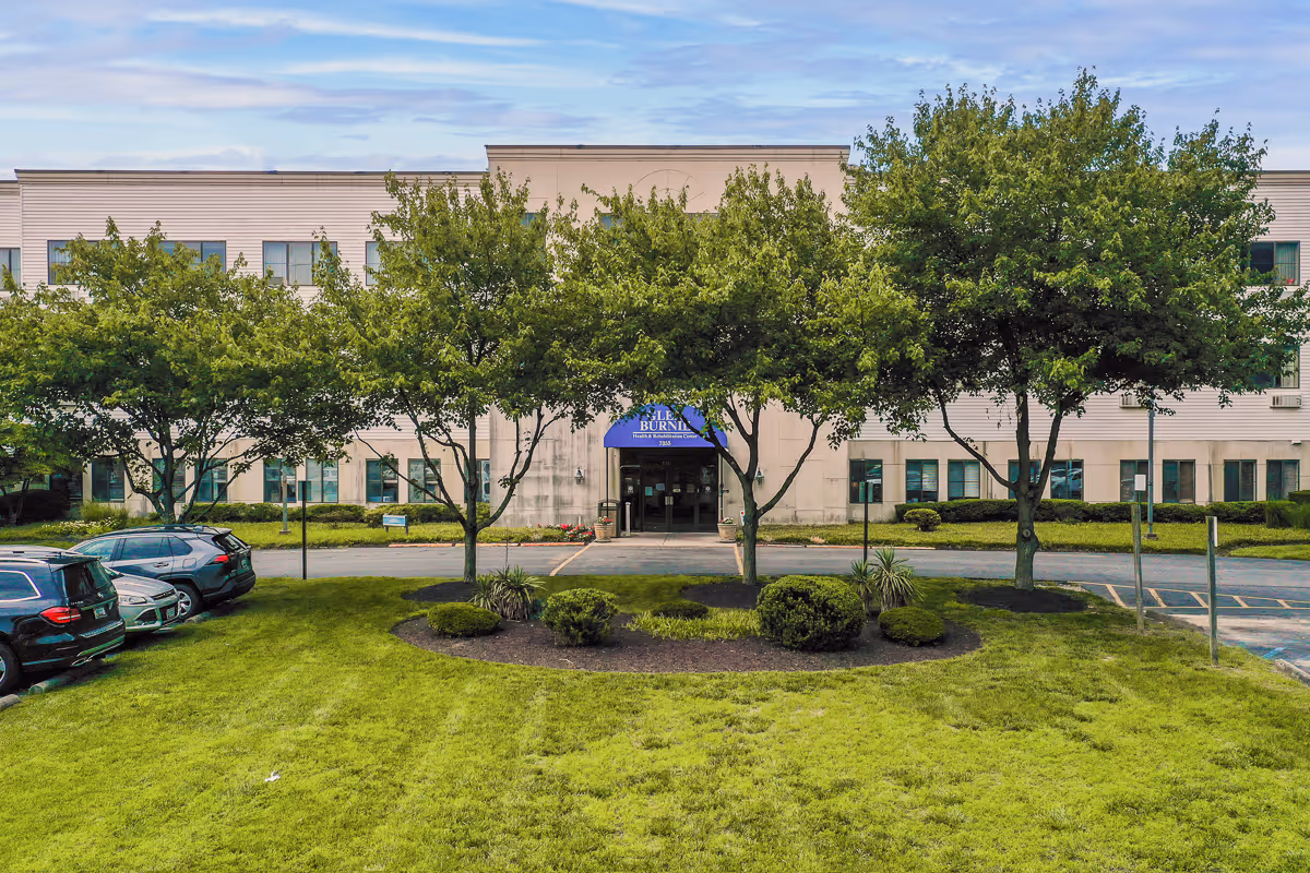 Front exterior view of Autumn Lake Healthcare at Glen Burnie building with a manicured lawn, several trees, parked cars on the left, and a blue awning above the entrance.