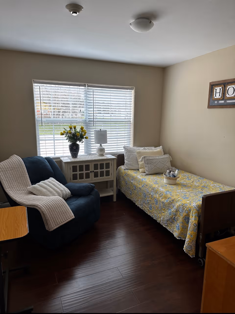Small senior living bedroom featuring a twin bed with a yellow floral quilt, a blue recliner with a blanket, and a window-side cabinet with a lamp and vase of flowers.