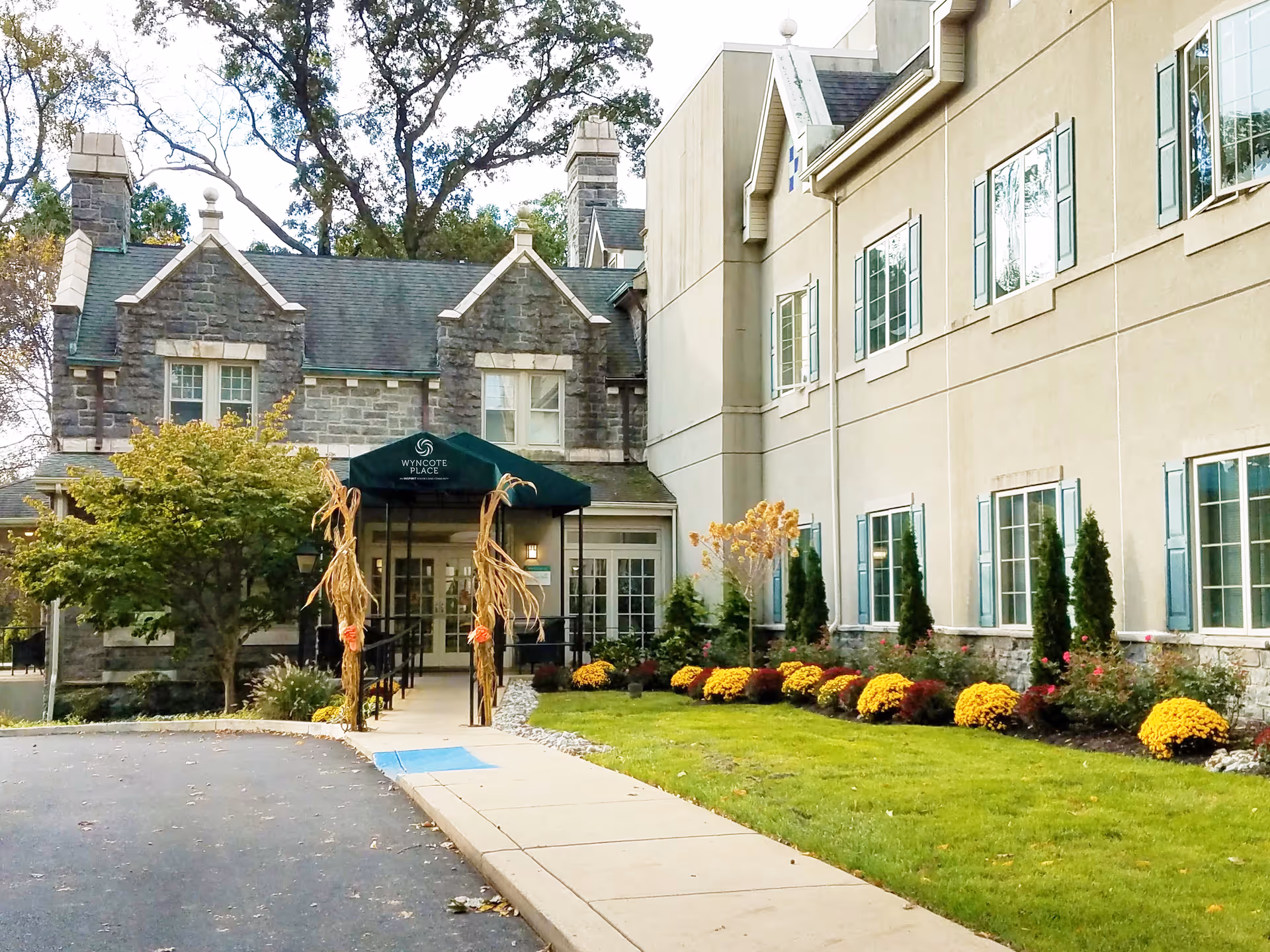 Exterior view of Wyncote Place, showing a building with stone and beige walls, multiple windows with blue shutters, a green awning over the entrance, and a landscaped garden with yellow flowers and small trees along the sidewalk.