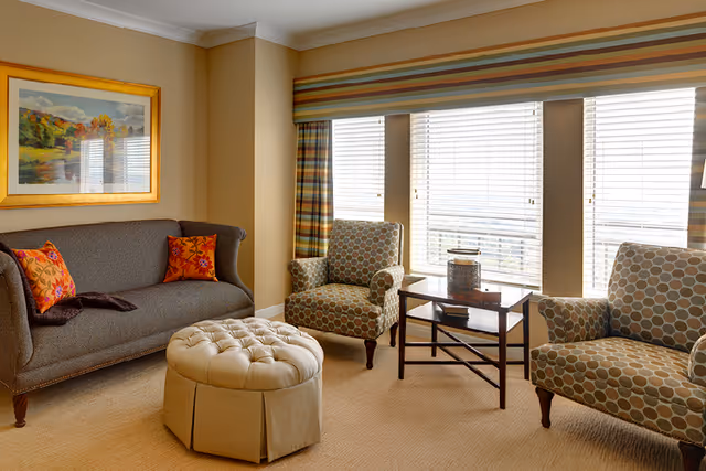 A cozy living room with a gray sofa adorned with two orange floral pillows, two patterned armchairs, a round tufted ottoman, and a wooden side table placed in front of three large windows with striped valances and blinds. A framed landscape painting hangs on the beige wall above the sofa.