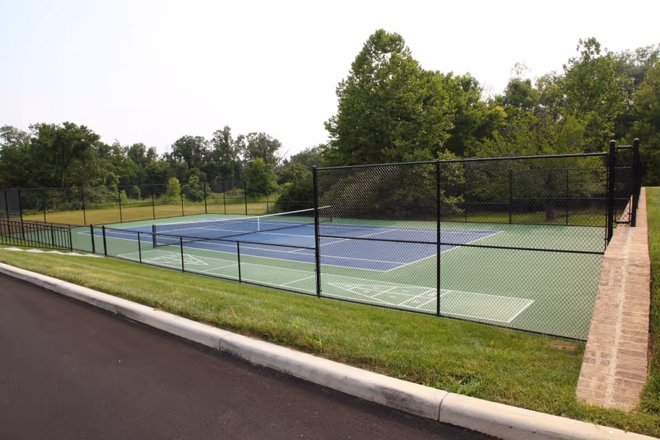 Outdoor tennis court surrounded by a black chain-link fence with green grass and trees in the background under a clear sky.