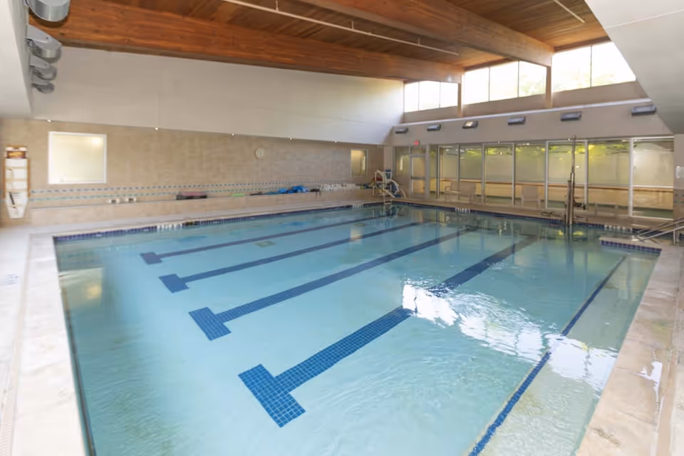 Indoor swimming pool with clear water, marked lanes, and a tiled deck. The pool area has large windows near the ceiling allowing natural light to enter, wooden beams on the ceiling, and a glass wall with doors on one side. Pool equipment and seating are visible along the far wall.
