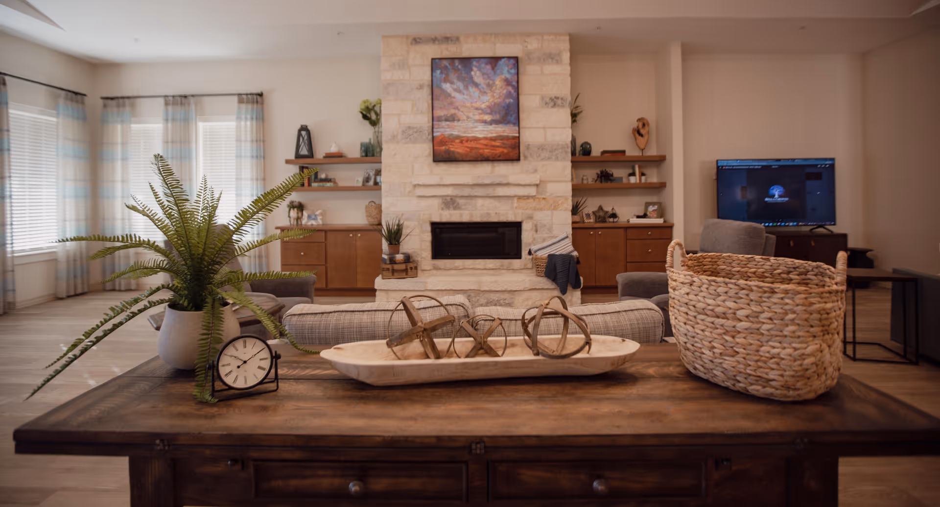 A cozy living room with a stone fireplace in the center, flanked by wooden cabinets and shelves with decorative items. In front of the fireplace is a wooden table with a potted fern, a small clock, a decorative tray with spherical objects, and a woven basket. There is a TV on the right side and large windows with light curtains on the left, allowing natural light to fill the room.