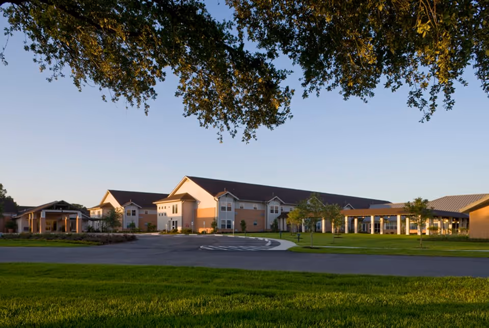 Exterior view of a large senior living facility building with a well-maintained lawn and trees in the foreground, under a clear sky during daylight.
