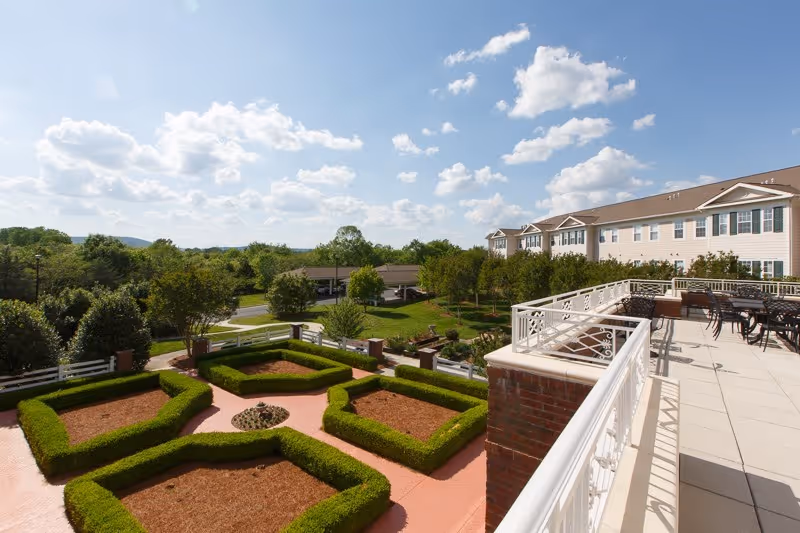 View from a balcony overlooking a landscaped garden with neatly trimmed hedges and pathways, adjacent to a large multi-story building under a partly cloudy sky.