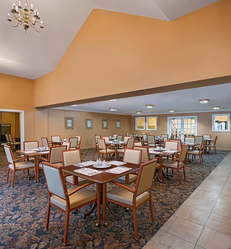 A spacious dining room with multiple square wooden tables, each set with placemats, napkins, and condiments. The chairs have wooden frames with beige cushioned seats and backs. The room has beige walls, a patterned carpet, and a tiled walkway on the right side. There are framed pictures on the wall and ceiling light fixtures providing illumination.