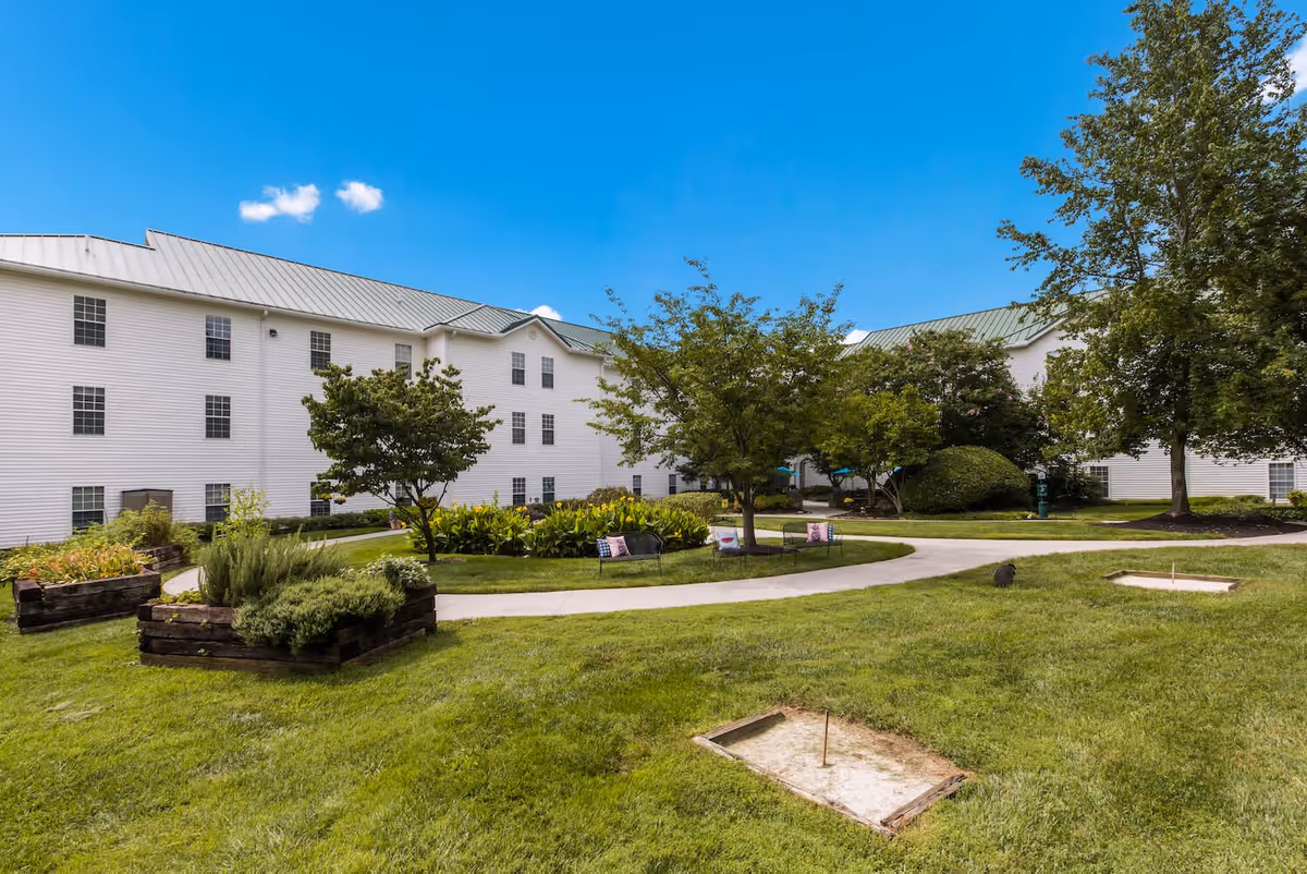 Outdoor garden area at Arbor Terrace Knoxville featuring green grass, trees, raised garden beds, benches with pillows, and a white multi-story building with a metal roof in the background under a clear blue sky.
