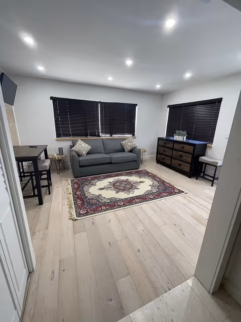 A modern living room with light wood flooring, a gray sofa with two patterned cushions, a decorative area rug with red and cream colors, a black dresser with six drawers, two small side tables, a small dining table with two chairs, and windows with dark blinds.